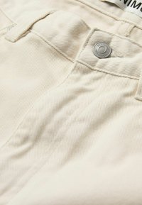 Pantalons en coton beige avec une surface texturée, dotés d'un bouton en métal argenté à la taille et d'un détail de poche cousue.