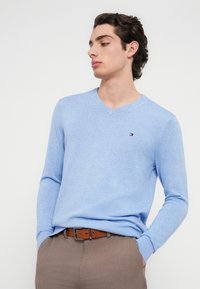 Φωτεινό γαλάζιο πουλόβερ με V-λαιμόκοψη από απαλό ύφασμα, με μακρύ μανίκι και μικρό λογότυπο στο στήθος. Συνδυάζεται με καφέ παντελόνια.