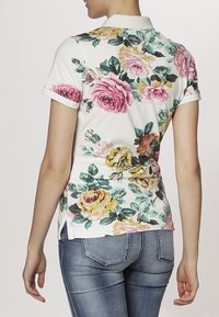 Chemise polo florale avec une base blanche ornée de grandes roses roses et jaunes, avec des manches courtes et un col. Design légèrement ajusté avec des fentes sur les côtés.