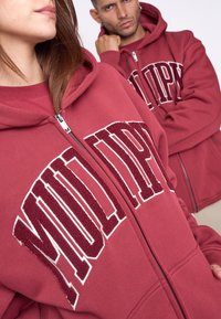 Due persone indossano felpe con zip in color bordeaux, decorate con un logo testurizzato bianco e burgundy. Le felpe sono dotate di tasche anteriori e vestibilità standard.