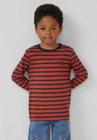 Jonge jongen met krullend haar, gekleed in een rood-en-marineblauw gestreept shirt met lange mouwen en een spijkerbroek met patches, staande met zijn handen achter zijn rug.