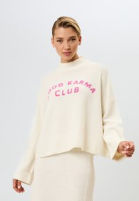 Crèmekleurige gebreide trui met een hoge hals, een ontspannen pasvorm en oversized mouwen; bevat "GOOD KARMA CLUB" in roze letters.