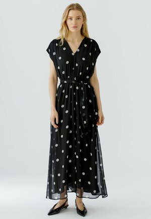 Femme blonde portant une robe maxi noire sans manches à pois blancs, une ceinture à la taille et des chaussures plates noires pointues, debout devant un fond uni.