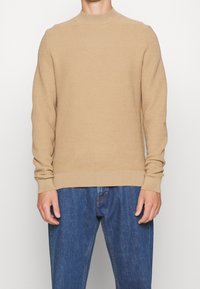 Beige gestrickter Pullover mit hohem Kragen, gerippten Bündchen und Saum. Getragen über blauen Jeans. Die Textur wirkt wie ein Wabenmuster.