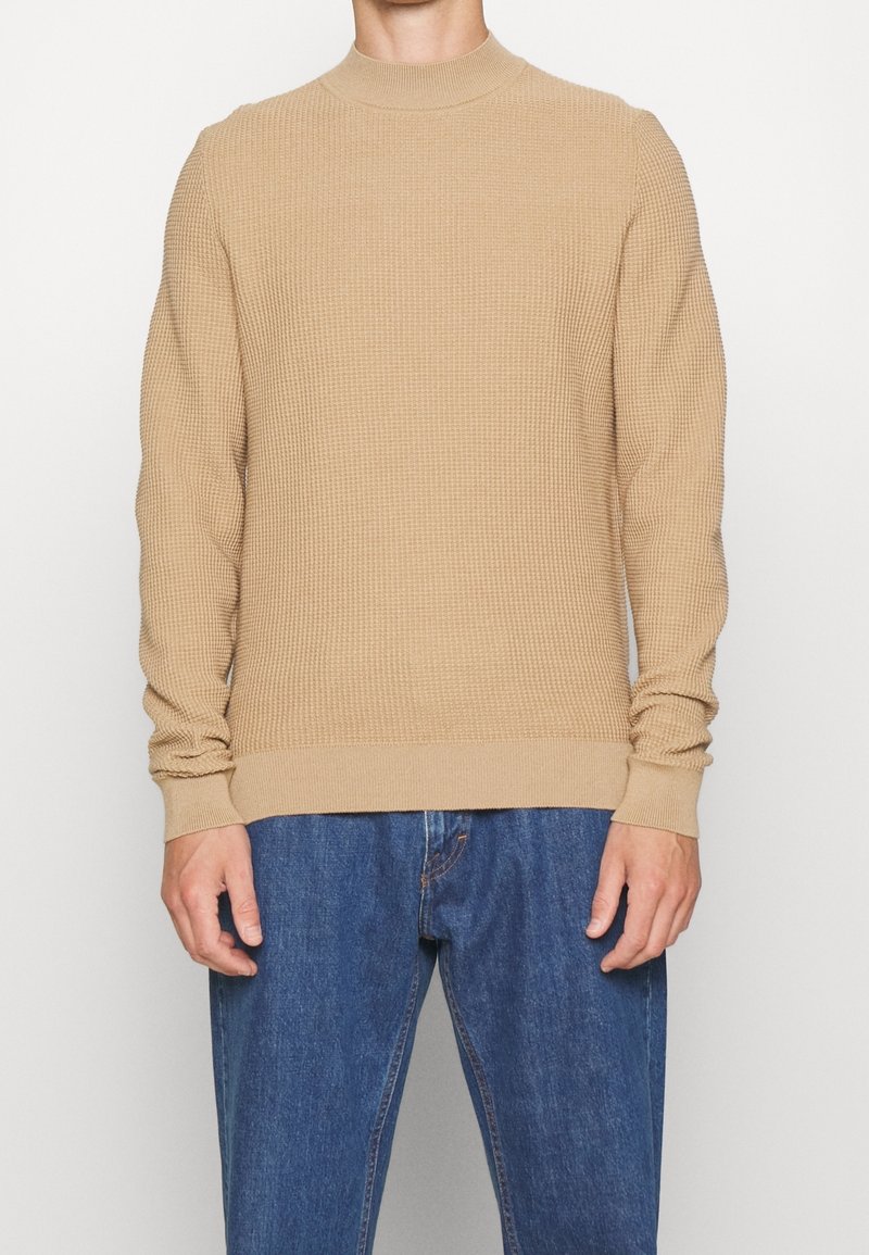 Beige gestrickter Pullover mit hohem Kragen, gerippten Bündchen und Saum. Getragen über blauen Jeans. Die Textur wirkt wie ein Wabenmuster.