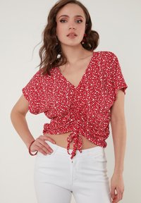 Rød blomster-bluse med korte ærmer, v-hals og samlingsdetaljer ved taljen. Kombineret med hvide jeans for kontrast.
