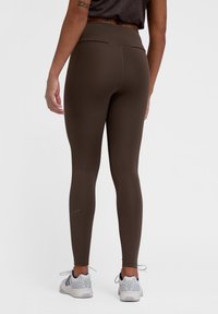 Bruna högmidjade leggings, slät textur, figurnära design, med en subtil gyllene detalj vid fotleden, tillsammans med grå sportskor.