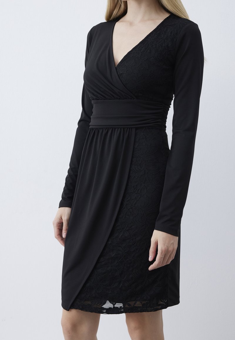 Femme portant une robe portefeuille noire à manches longues avec un détail en dentelle sur un côté et une taille ajustée, debout devant un fond clair uni.