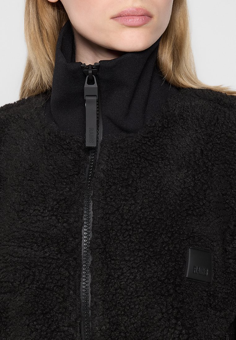Veste polaire noire avec col haut, dotée d'une fermeture éclair sur le devant et d'un tissu côtelé. Apparence texturée et douce avec une écusson logo sur la poitrine.