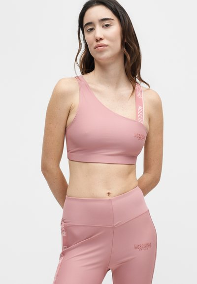 Γυναίκα που φοράει ροζ αθλητικό σουτιέν με έναν ώμο και αντιστοιχισμένα κολάν με το λογότυπο Moschino Active στο λουρί και στο μηρό.