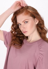 Top scurt de culoare mauve deschis, cu detalii pliate pe lateral, textură moale, guler rotund și croială lejeră. Modelul pozează cu o mână ridicată.