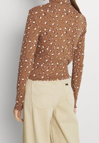 Braunes langärmliges Oberteil mit weißem und rotem Blumenmuster, gerippter Textur und gewelltem Saum, kombiniert mit beigen, weit geschnittenen Hosen mit aufgesetzten Gesäßtaschen.