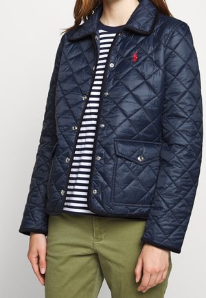 Žena v tmavě modré prošívané bundě se zapínáním na patentky, pruhovaném černo-bílém tričku a olivově zelených kalhotách, stojící na jednobarevném pozadí.