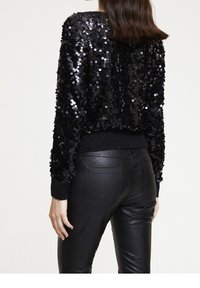 Svart paljettjacka med en kortad design, avslappnad passform och ribbade muddar. Bärs över svarta läderbyxor som visar upp en glänsande textur och intrikata detaljer.
