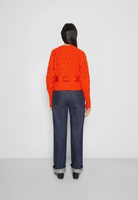 Pull en maille orange vif avec un motif en diamant texturé et des poignets côtelés, associé à un jean large bleu foncé avec des ourlets retroussés.