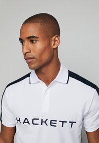 Polo bianco con colletto e dettagli sulle spalle in blu navy, grande logo "HACKETT" in blu navy, maniche corte e vestibilità classica.