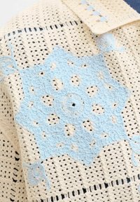 Kremfarget heklet stoff med et blått stjerneformet heklet motiv overlagt, som viser detaljert håndlaget tekstilarbeid.