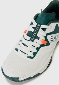 Pantofii sport albi cu suprafață texturată, accente verzi și un sistem de prindere pentru șireturi de culoare portocalie. Au șireturi albe și un model specific pe talpă.