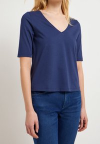 Námořnická modrá v-neck tričko s krátkými rukávy, vyrobené z měkké tkaniny. Má uvolněný střih a rovný lem. Kombinováno s tmavými džínami.