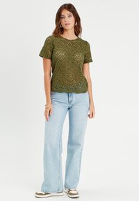 Top en crochet vert olive à manches courtes, présentant des motifs complexes et un ourlet festonné, associé à un jean bleu clair évasé.