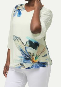 Blusa leggera e fluida con scollatura a V, maniche 3/4 e stampa floreale nelle tonalità di blu e beige su sfondo bianco.