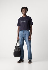 Jūrų mėlynos medvilnės marškinėliai su "BOSS" logotipu, derinami su šviesiai mėlynomis tiesiomis kelnėmis. Modelis laiko juodą maišelį, vilkėdamas tamsius sportbačius.
