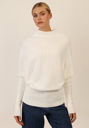Weißer, gerippter Pullover mit hohem Kragen und weiten Ärmeln. Kombiniert mit dunkelblauen Jeans mit dekorativen Nieten. Angenehme Textur.