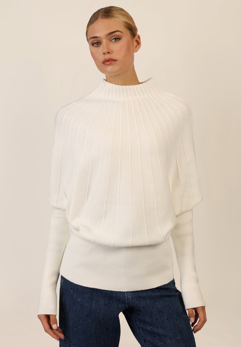 Weißer, gerippter Pullover mit hohem Kragen und weiten Ärmeln. Kombiniert mit dunkelblauen Jeans mit dekorativen Nieten. Angenehme Textur.