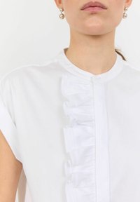 Camicia bianca a colletto con maniche corte, caratterizzata da un bordo arricciato verticale sul davanti e un tessuto morbido.