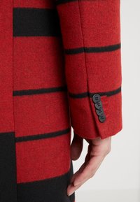 Czerwono-czarny pasiasty rękaw płaszcza z wełny, z widocznymi guzikami i detalami szwów. Tkanina wygląda na teksturowaną i ciepłą.