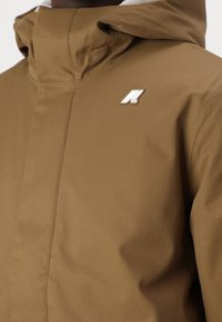 Giacca con cappuccio marrone realizzata in tessuto liscio. Presenta una chiusura con zip, dettagli delle cuciture e un logo bianco sulla parte superiore sinistra del petto.