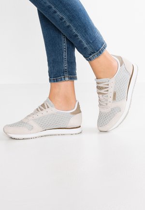 Fødder iført lyse beige mesh-sneakers med hvide såler, kombineret med opsmøgede blå jeans.