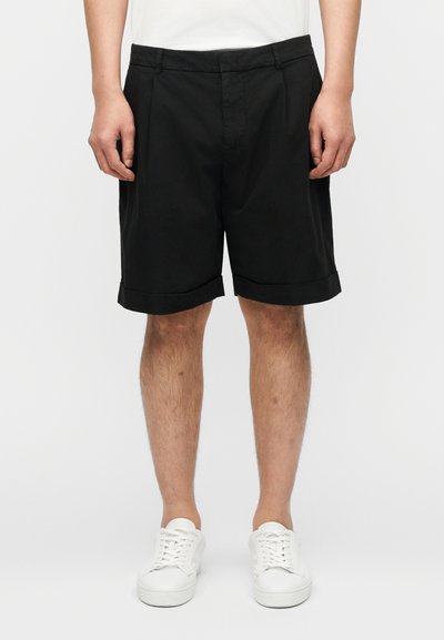 Homme portant un short noir jusqu'aux genoux, des baskets blanches et une chemise blanche, debout devant un fond clair uni.