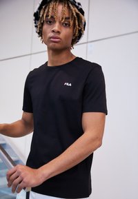 Camiseta negra de manga corta con cuello redondo y un pequeño logo de FILA en blanco y rojo en el lado izquierdo del pecho. Tela de algodón suave.
