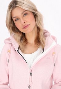 Blonde woman with wavy hair wearing a light pink zip-up hoodie over a white shirt, looking slightly tilted with a soft expression.