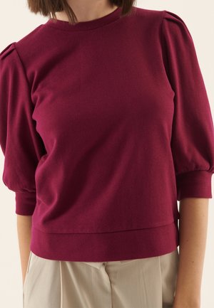 Vrouw draagt een nauwsluitende donkerrode top met opgeblazen mouwen tot de ellenboog en beige, hooggetailleerde, geplooide broek.