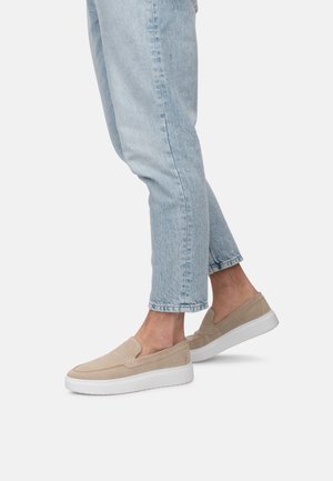 Zapatos slip-on de ante beige con una suela de goma blanca, con un diseño minimalista y sin cordones visibles, llevados con unos vaqueros de mezclilla azul claro.