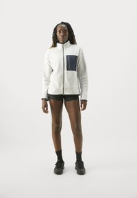 Grau Fleecejacke mit einer marineblauen Brusttasche und schwarzem Reißverschluss, ausgestattet mit einem hohen Kragen und langen Ärmeln, kombiniert mit schwarzen Shorts und Sportschuhen.
