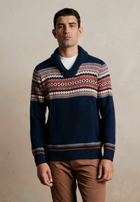 Granatowy sweter dzianinowy z kołnierzem szalowym, z czerwono-kremowymi wzorami geometrycznymi na klatce piersiowej. Dziane mankiety i dół.