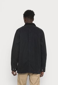 Chaqueta negra con un cuello de pana, de manga larga y dos puños abotonados. Textura suave con un corte recto, vista desde la parte posterior.