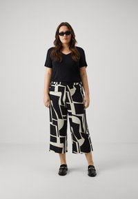 Un haut noir à manches courtes en V, un pantalon large noir et crème à motifs, et des mocassins noirs avec des détails en chaîne dorée, portés avec des lunettes de soleil noires.