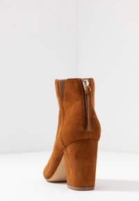 Ruskea mokkanahkainen nilkkabooti, jossa on paksu korkea korko ja takana vetoketj closure, aseteltuna valkoista kulmaa vasten.