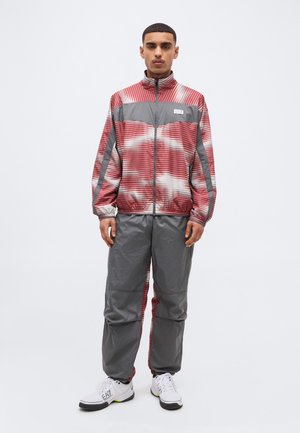 Άνδρας που στέκεται φορώντας μια κόκκινη και γκρι ριγωτή ζακέτα με φερμουάρ, γκρι παντελόνι και λευκά αθλητικά παπούτσια με μαύρη σόλα σε λευκό φόντο.