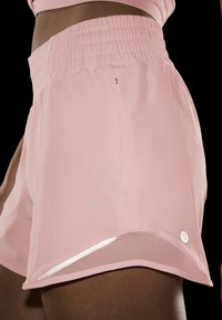 Suur lähedus inimesest, kes kannab heledat roosa värvi spordipükse, millel on rüüstatud vöökoht, külgmine tõmblukktasku, võrkpits ja Lululemoni logo mustal taustal.