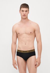 Schwarze Slips mit einem braunen Gummibund, der das "HUGO"-Logo trägt. Der Stoff erscheint glatt und hat ein eng anliegendes Design.