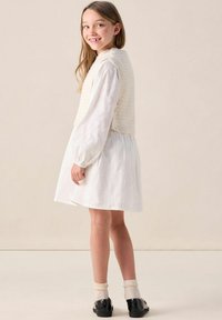 Cremfarbener, gestrickter Weste über einem weißen Langarmkleid aus strukturiertem Stoff. Schwarze Schuhe und hellfarbige Socken runden das Outfit ab.