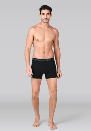 Mand med kort mørkt hår står barbent iført sorte boxershorts med "MUCHACHOMALO" taljebånd på grå baggrund.