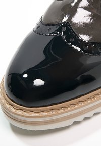 Glansig svart läderbrogue-sko med beige sytt sula och vit slitbanan, närbild av tå och övre detaljer.