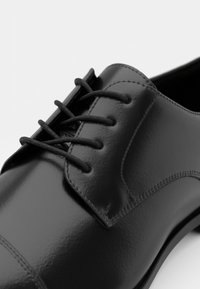 Zapato de vestir de cuero negro con textura suave, que presenta un sistema de cordones de 6 ojetes y una puntera redondeada. Detalle de costura sutil en la parte superior.