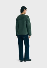 Giacca verde trapuntata con taglio dritto e spalle larghe. Abbinata a pantaloni in denim scuro e stivali neri alla caviglia. Design semplice con cuciture verticali.